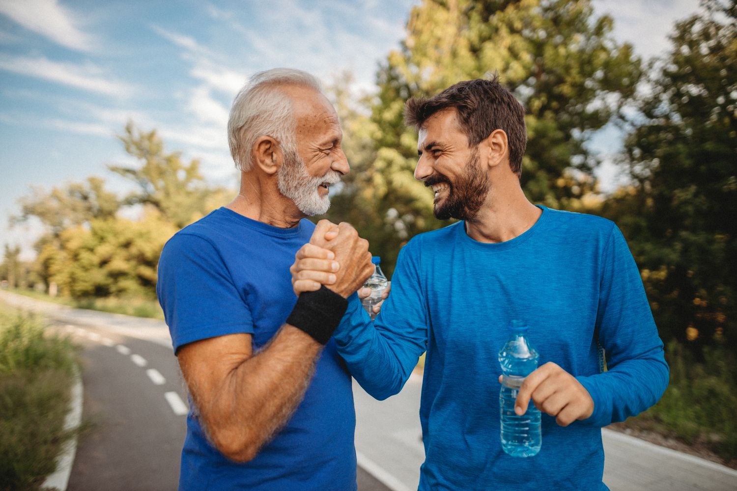 An older man with a white beard and a younger man clasp hands in a celebratory handshake, smiling at each other while holding water bottles on an outdoor running track.